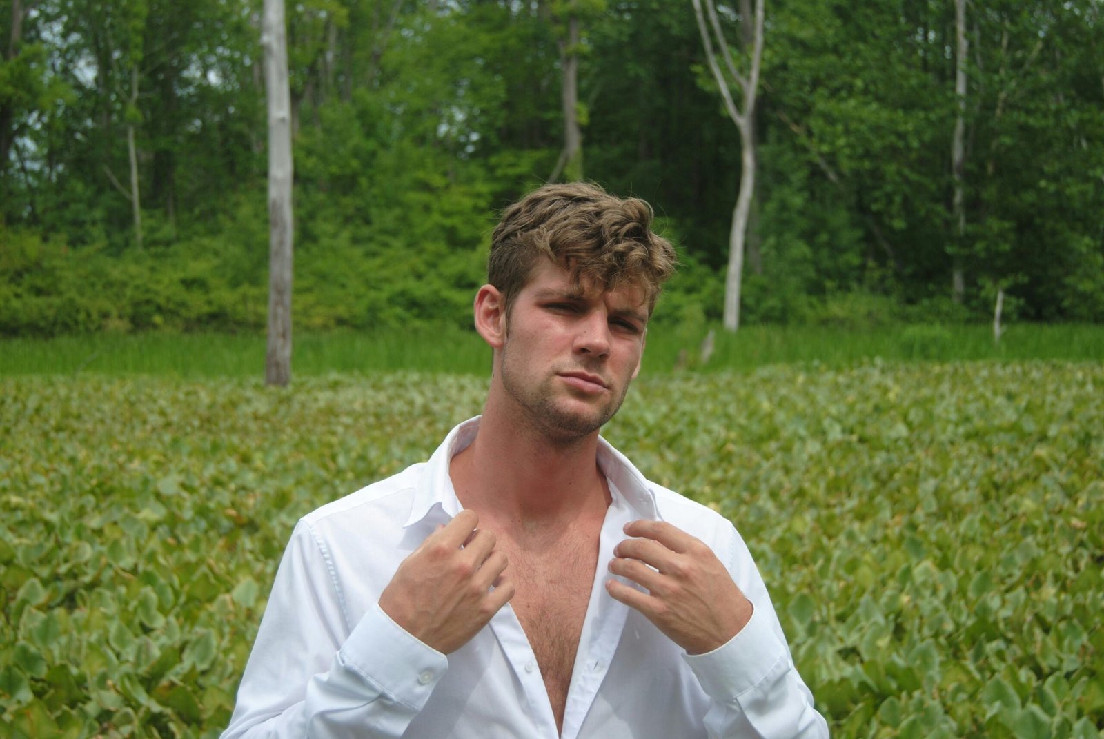 A young man in a white shirt poses confidently in a lush, green field.